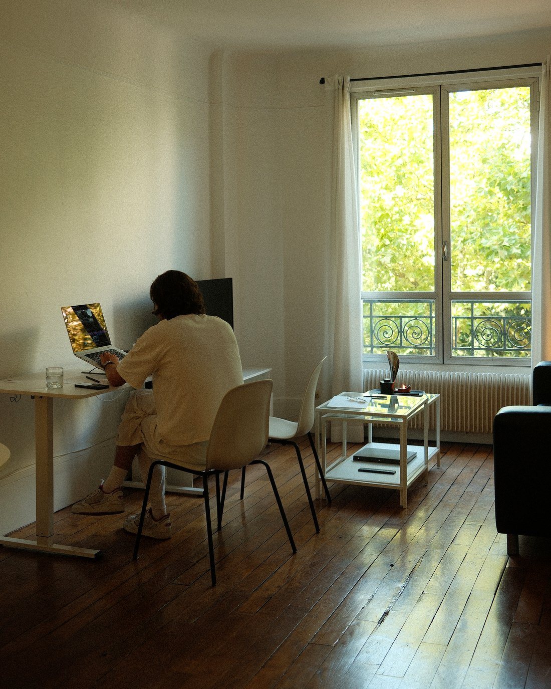 Valentin, de dos à son bureau devant un ordinateur portable, dans une pièce claire ouverte sur une fenêtre et des arbres.